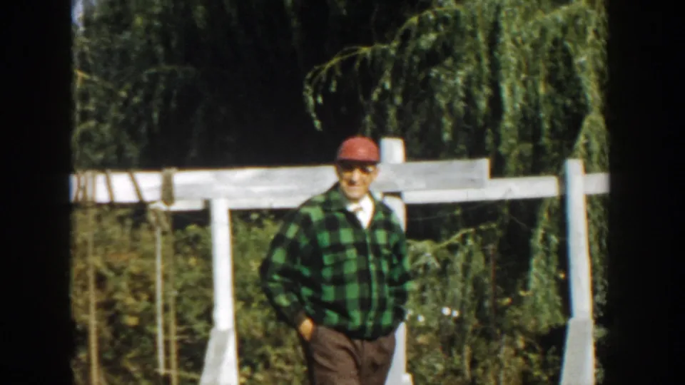 Adult man in red cap and plaid shirt stands with hands in pockets by a white fence in 1958 Michigan.