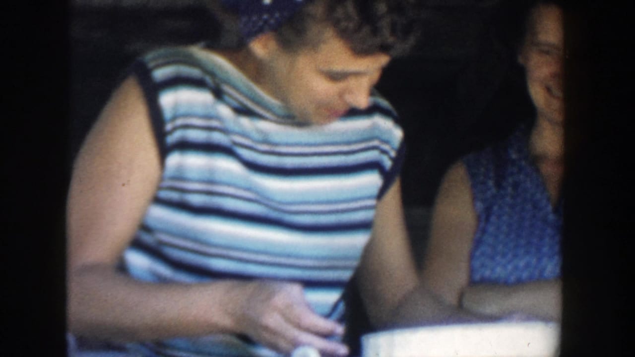 In 1958 Michigan home-movie footage, a woman in a striped sleeveless top looks down as another woman stands behind her in a dim interior.