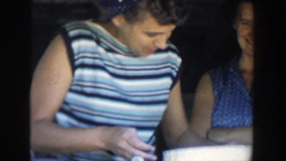 In 1958 Michigan home-movie footage, a woman in a striped sleeveless top looks down as another woman stands behind her in a dim interior.
