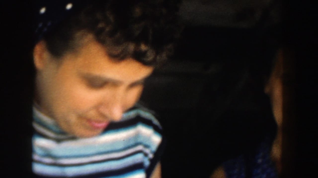 A woman in a striped sleeveless top looks downward in a dim 1958 Michigan home-movie interior, with another figure at the edge.