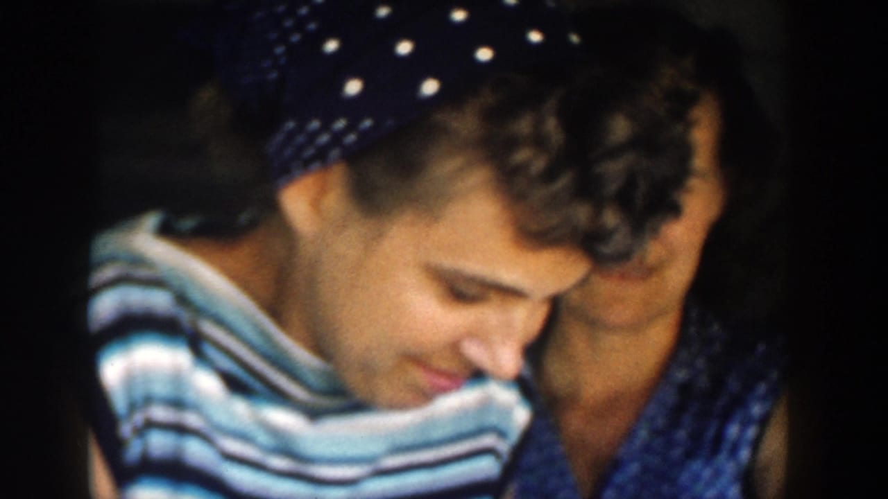 Two women appear indoors in Michigan home-movie footage from 1958, one leaning forward in stripes and one smiling behind in a blue patterned dress.