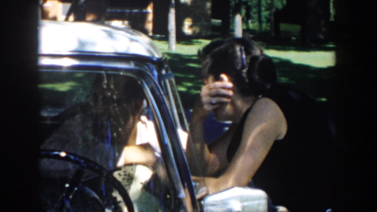 In 1958 Michigan home-movie footage, a woman in a dark sleeveless dress leans toward a car window as sunlight falls across the vehicle.