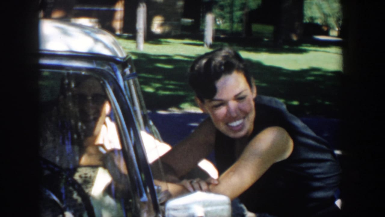 Smiling woman leans on a parked car and talks with someone inside.