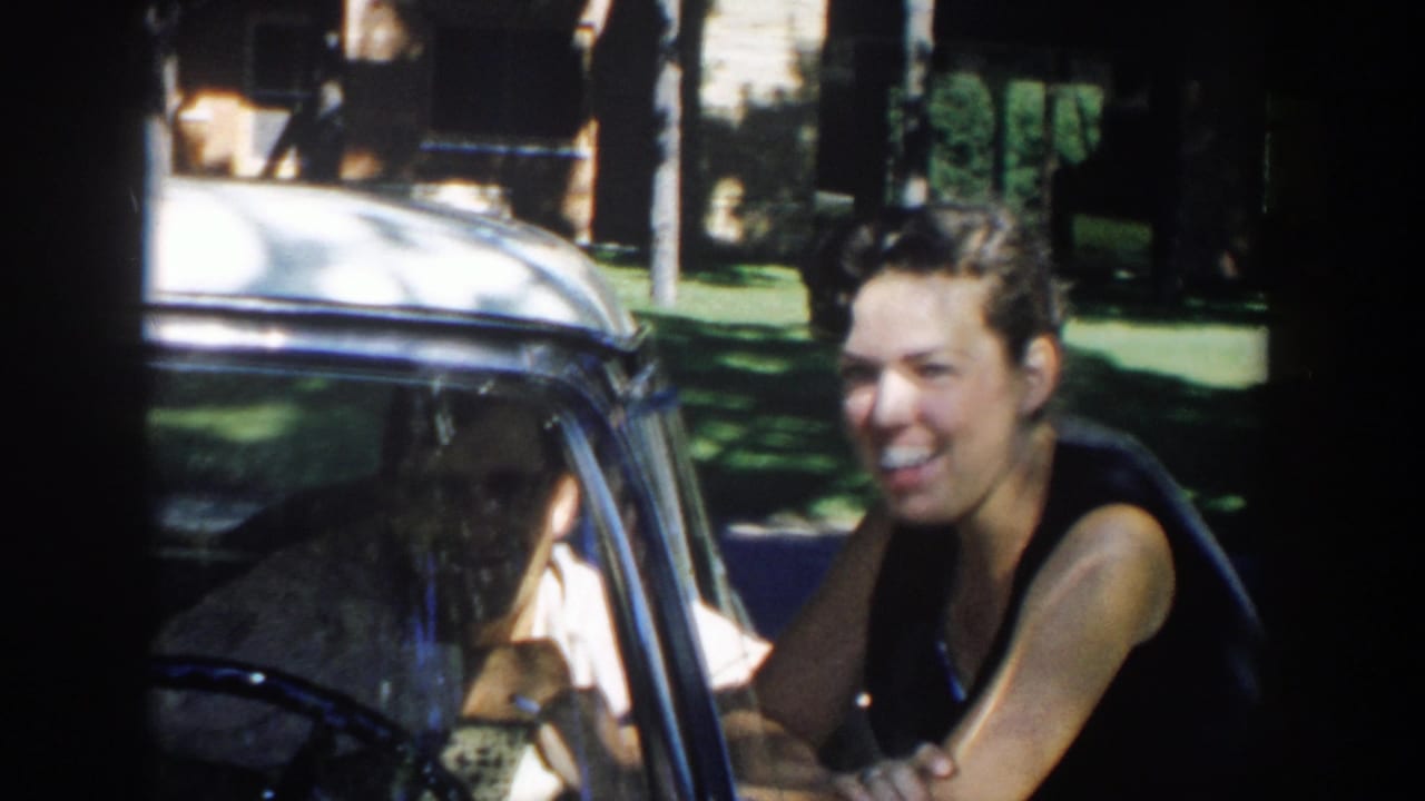A woman smiles beside a parked car in 1958 Michigan, with trees and lawn behind her and the driver partly seen through the glass.