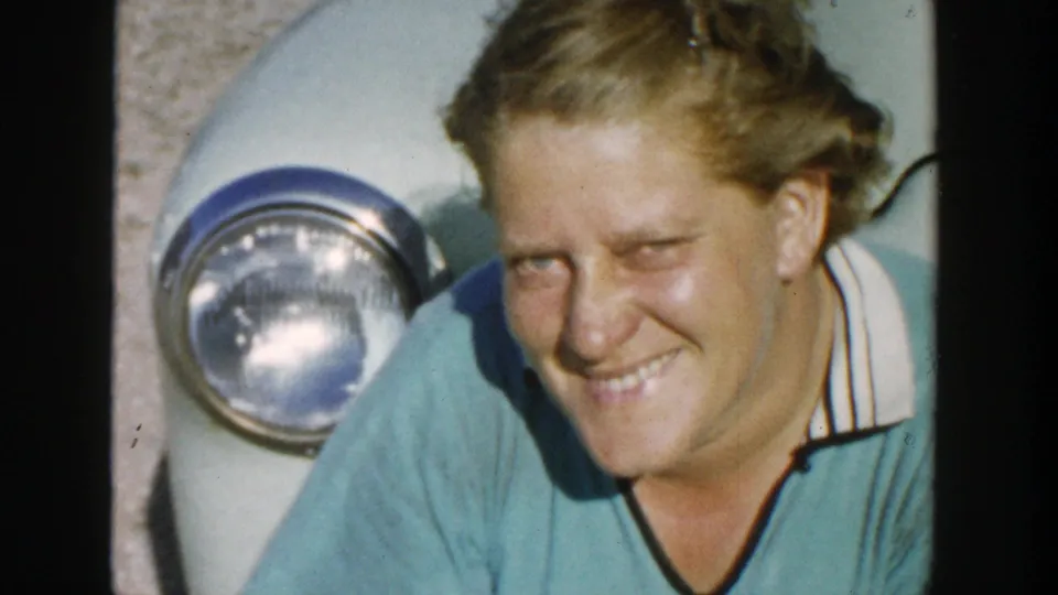 A blonde woman smiles toward the camera in bright sunlight beside a light-colored vintage car in 1958 Michigan footage.
