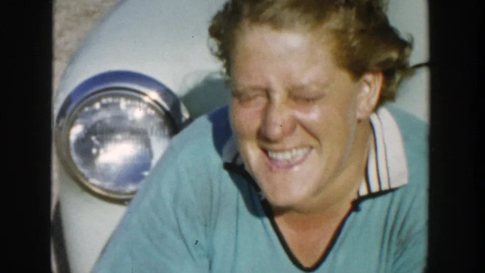 The woman closes her eyes in a wink or laugh while standing beside the light-colored car in the 1958 Michigan home movie.