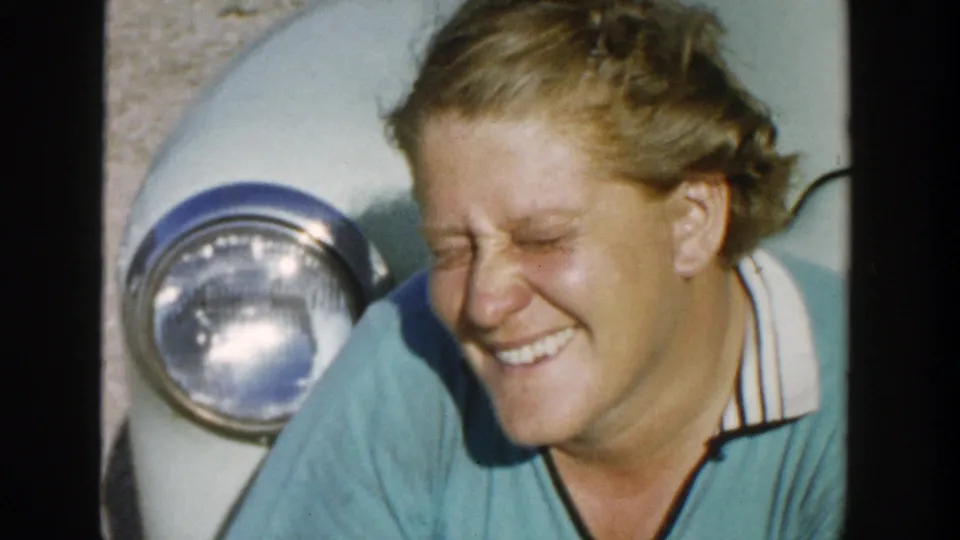 A blonde woman laughs with eyes closed beside a light-colored car, captured as candid 1958 Michigan 8mm home-movie footage.