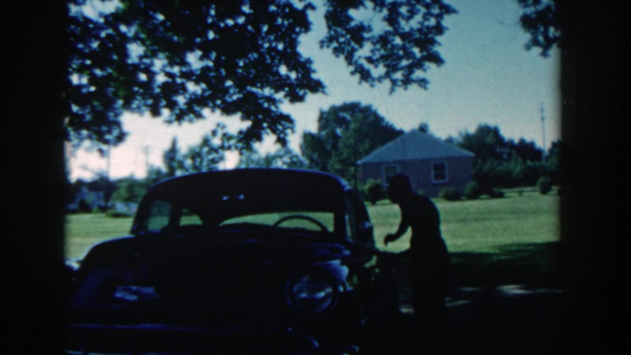 1958 Michigan home-movie frame with a silhouetted boy near a dark 1950s car, lawn, trees, and a small house beyond.
