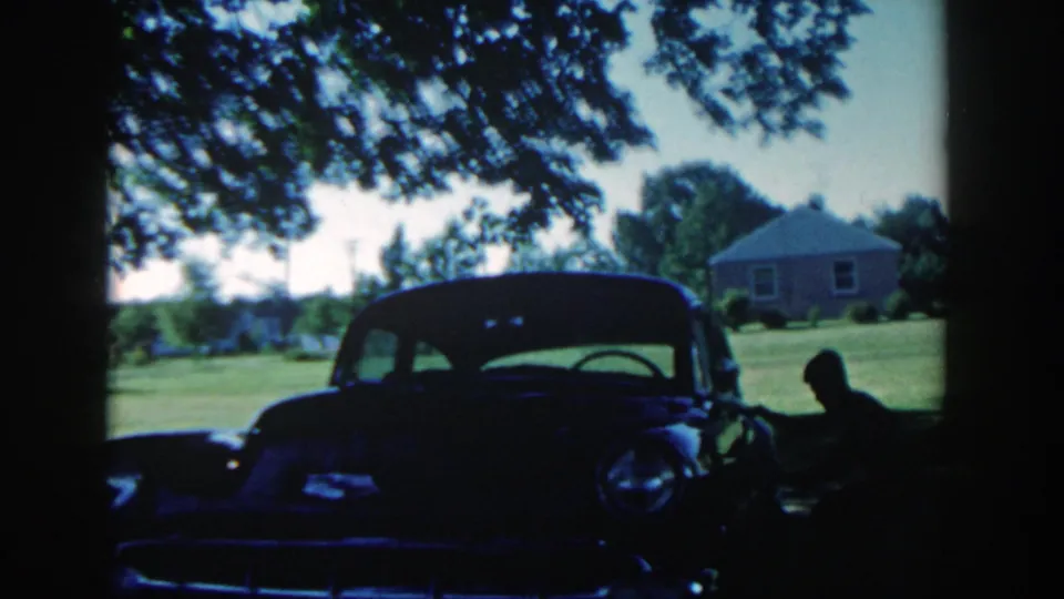The 1958 home-movie frame shows a boy crouched low beside the car, silhouetted against the sunny lawn and nearby house.