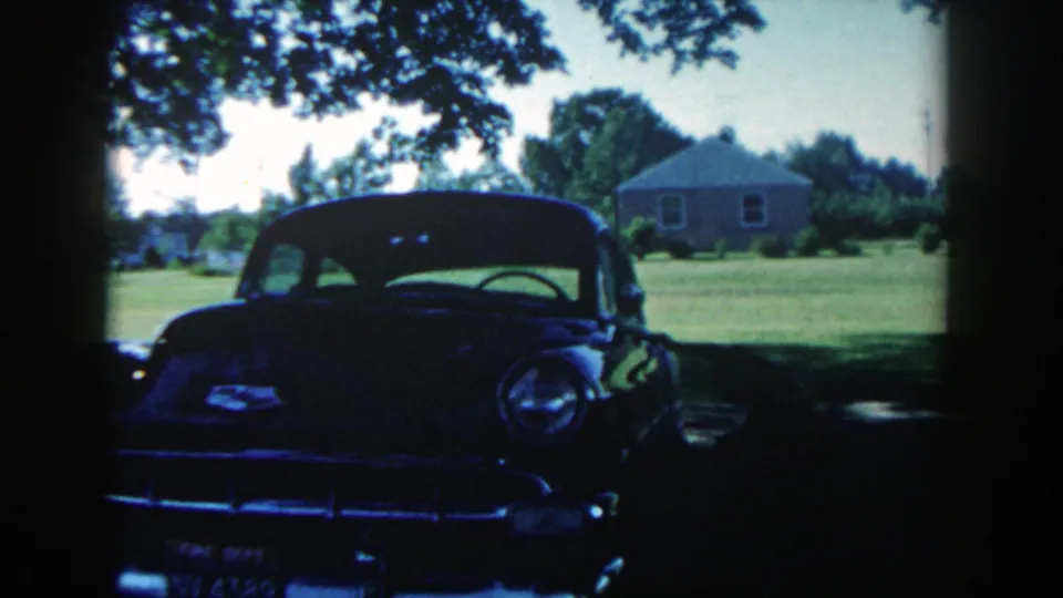 A dark 1950s-style car fills the shaded foreground while the bright Michigan yard and small house remain visible behind it.