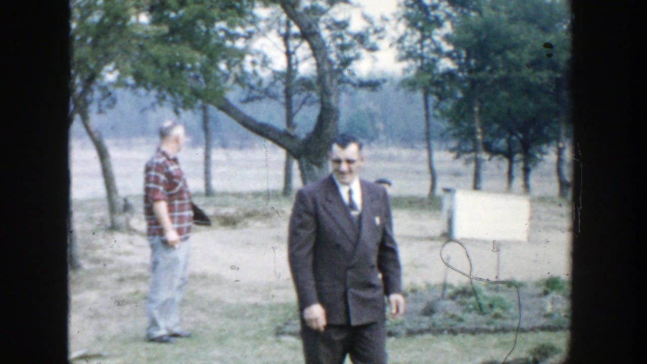 1958 Michigan 8mm frame showing a suited man walking outdoors, with a plaid-shirted man, trees, grass, and a small board behind him.