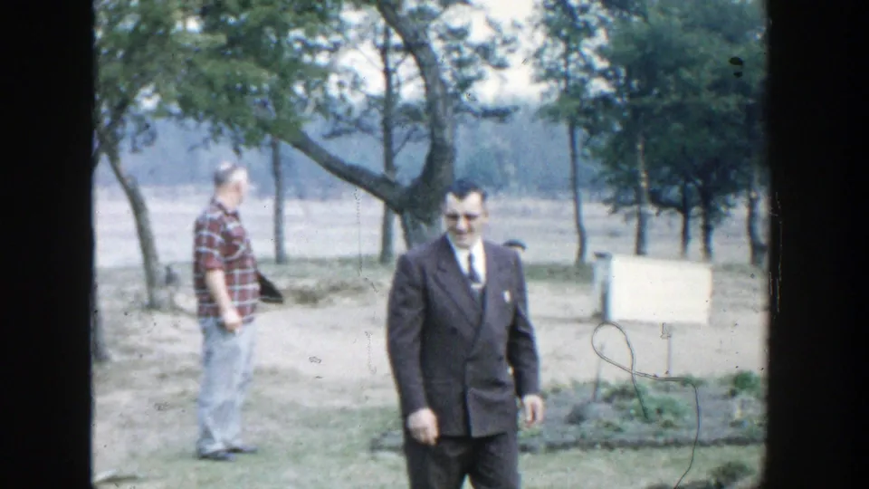 1958 Michigan 8mm frame showing a suited man walking outdoors, with a plaid-shirted man, trees, grass, and a small board behind him.