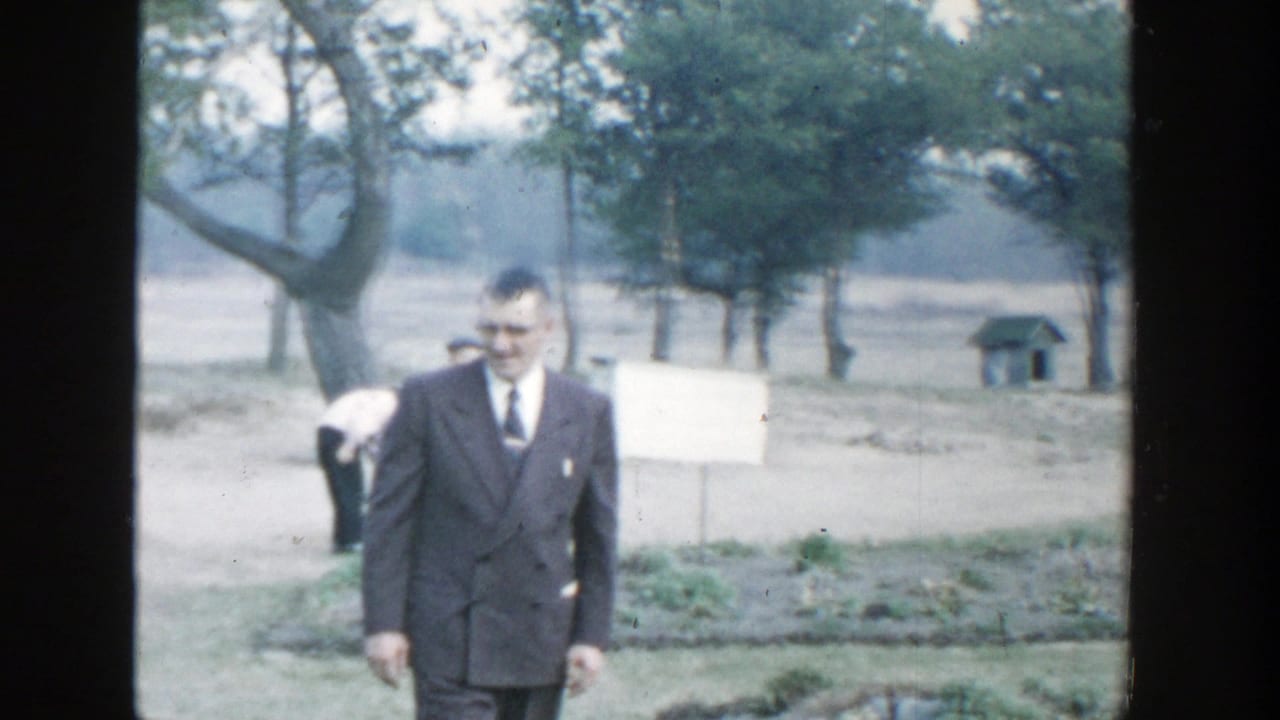 1958 Michigan home-movie frame of a man in a dark suit walking through a grassy field near trees and a pale sign or board.