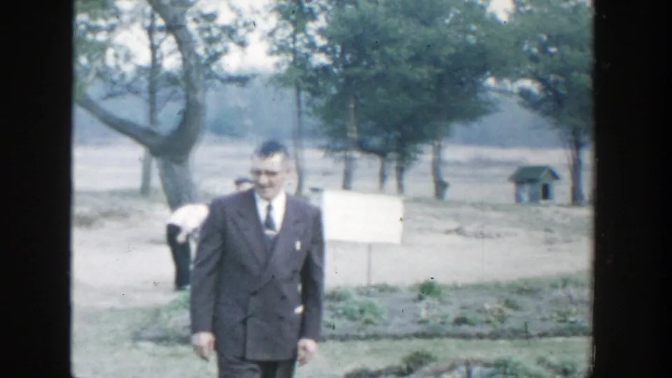 1958 Michigan home-movie frame of a man in a dark suit walking through a grassy field near trees and a pale sign or board.