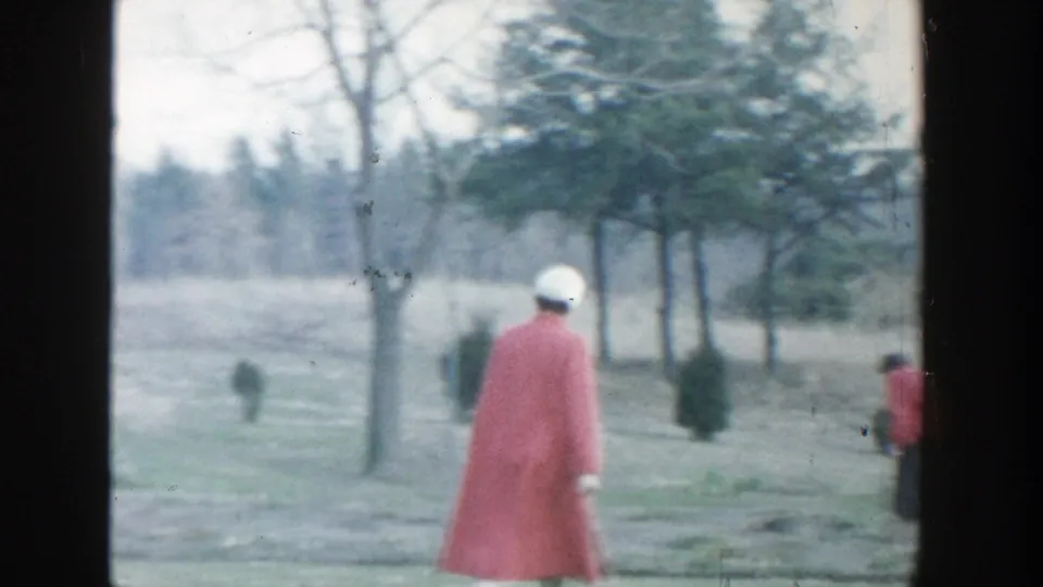1958 Michigan 8mm frame showing a woman in a red coat and white hat walking away through a grassy, tree-lined outdoor area.