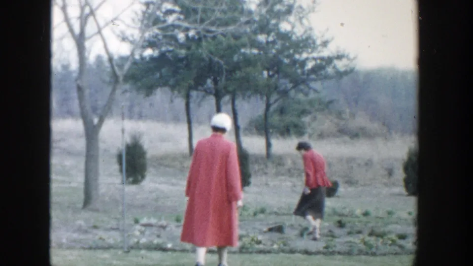 1958 Michigan home-movie frame with two women in red and pink coats walking through a grassy field near low garden beds and trees.