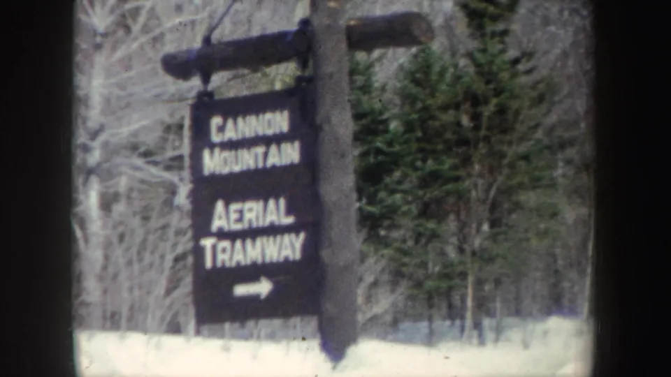 The sign for Cannon Mountain Aerial Tramway appears in front of evergreens and bare winter trees, with snow along the ground.