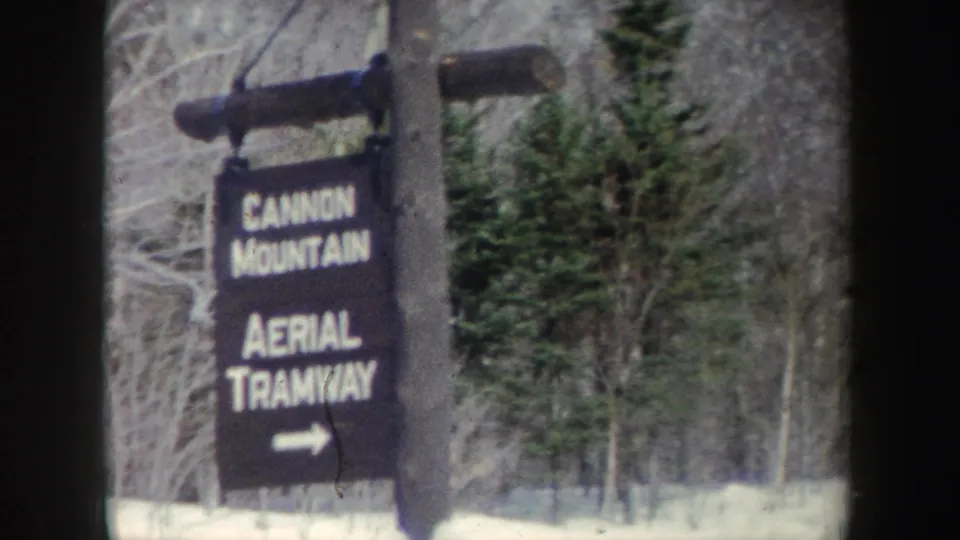 The black-and-white-lettered tramway sign is filmed from close range, with snow and forest visible behind it.