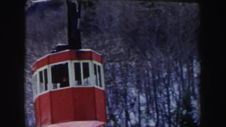 1962 home-movie frame showing a red gondola cabin on cable hardware, with a dark snowy forest behind it.