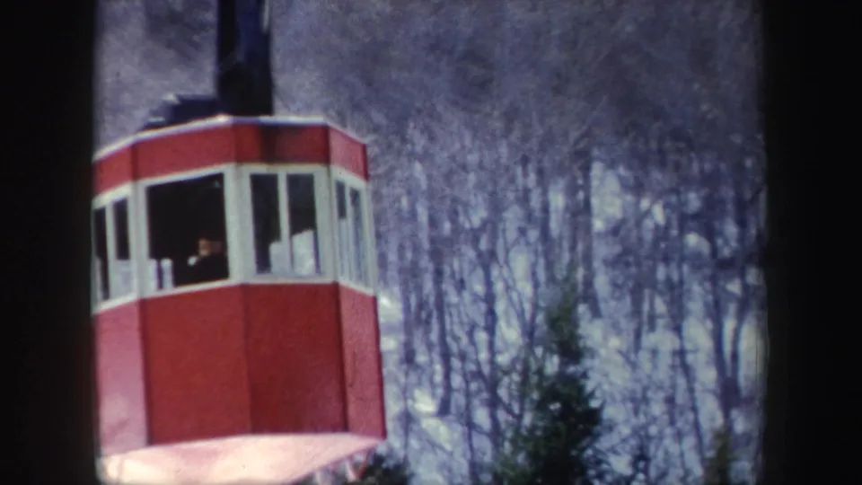 Vintage 8mm frame of a red aerial tramway gondola with a passenger visible through the window over a snowy wooded slope.