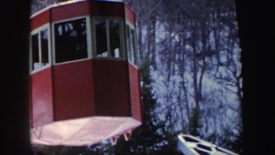 1962 New York frame with a red cable-car cabin seen close up, windows darkened by shadow and trees behind it.
