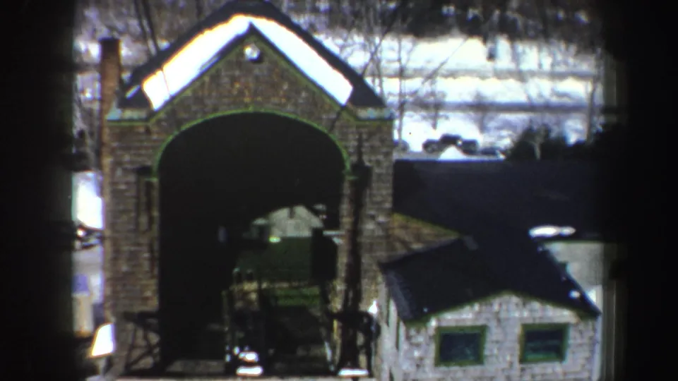 1962 New York home-movie frame widening to show the brick archway, snow beyond, parked cars, and a small structure at right.