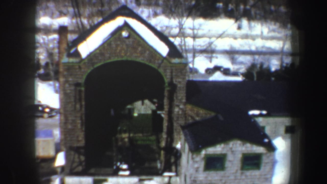 Snowy ski area seen from a lift, with a stone-faced structure, cars, buildings, and dark support framing.