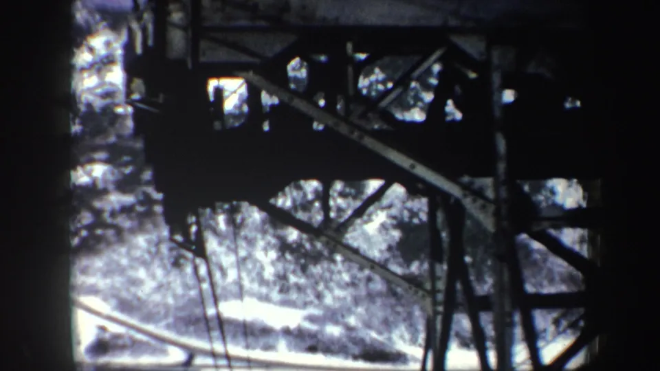 1962 New York ski lift POV with heavy metal support structure in the foreground and a snowy road or path below.