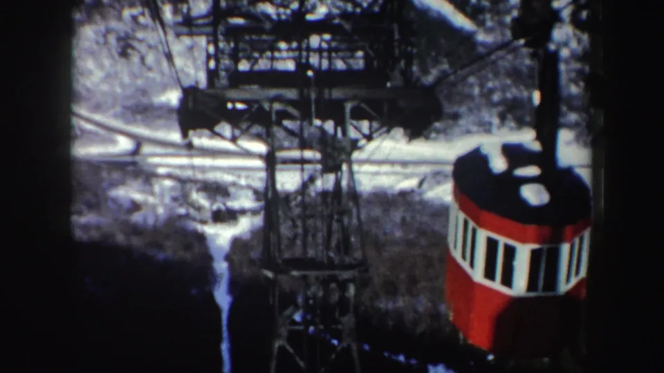 1962 New York home-movie frame with a red and white cable car cabin passing near a steel tram tower above snow-covered trees.
