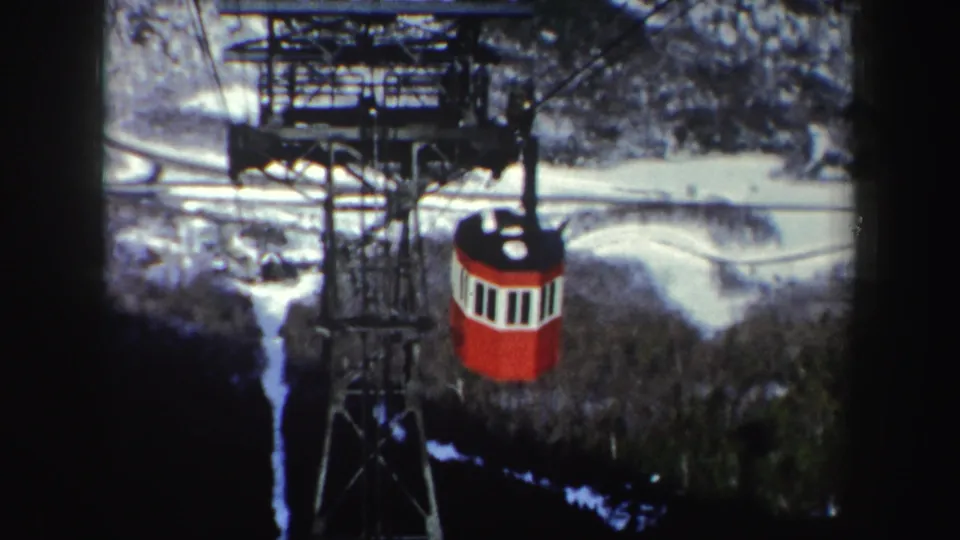 1962 8mm frame likely in New York showing a red aerial tram cabin beneath metal cable works above a snowy wooded slope.