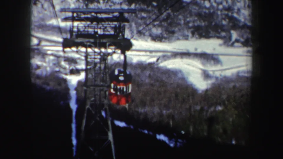 1962 New York frame showing the red tram cabin farther away along its cables, with steel supports and a snow-dusted forested hillside below.