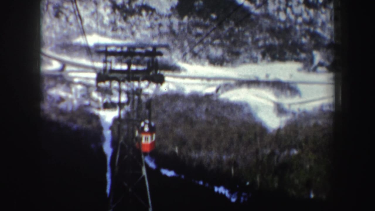 Small red cable car moving along cables with snowy landscape behind it.