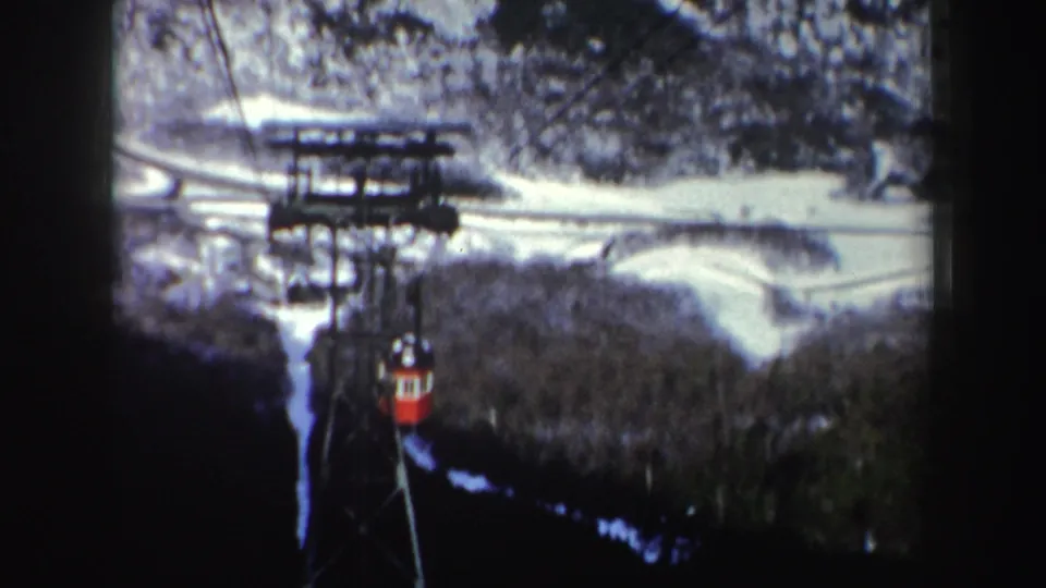 1962 New York home-movie frame of a red cable car descending across a snowy mountainside with trees below.