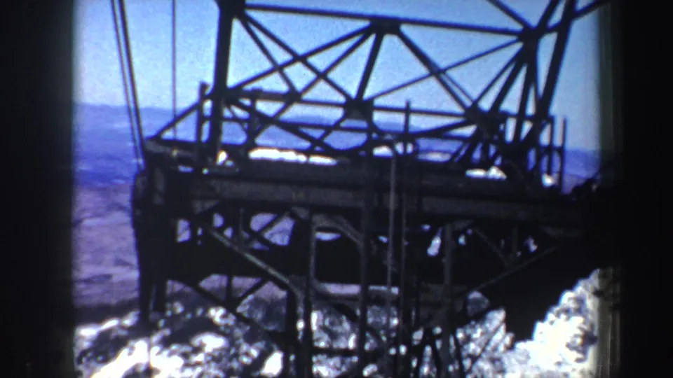 1962 New York cable-car ride frame showing trusswork, cables, and snowy ground with distant mountains beyond.