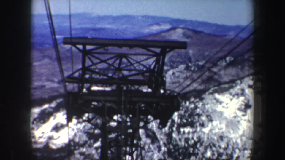 1962 New York home-movie frame with a cableway support tower and cables over rocky, snow-patched mountain slopes.