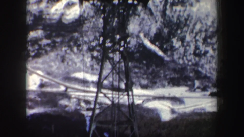 Passenger-view 8mm frame showing the lower section of a cableway tower with snow patches, trees, and a pale road or trail below.