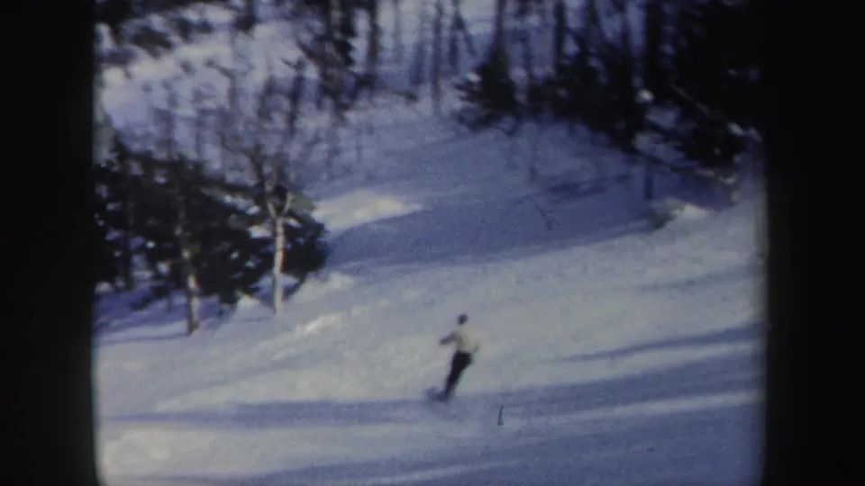 1962 New York 8mm frame showing a skier in a light jacket and dark pants carving across a snowy hillside.
