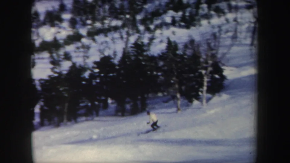 1962 New York home-movie frame of a skier turning low on a snowy run bordered by winter trees.