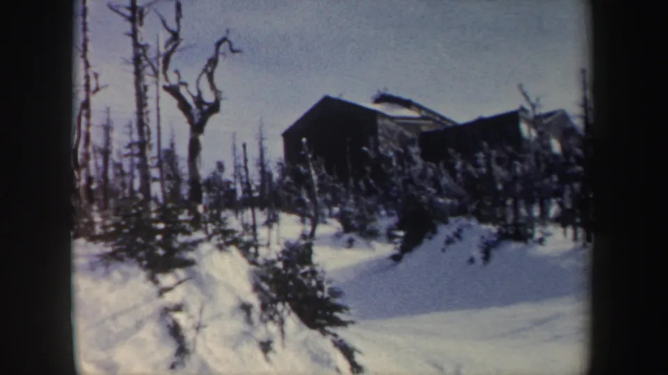 1962 New York 8mm frame with a dark cabin-like building above a snowbank, surrounded by bare trees and small evergreens.