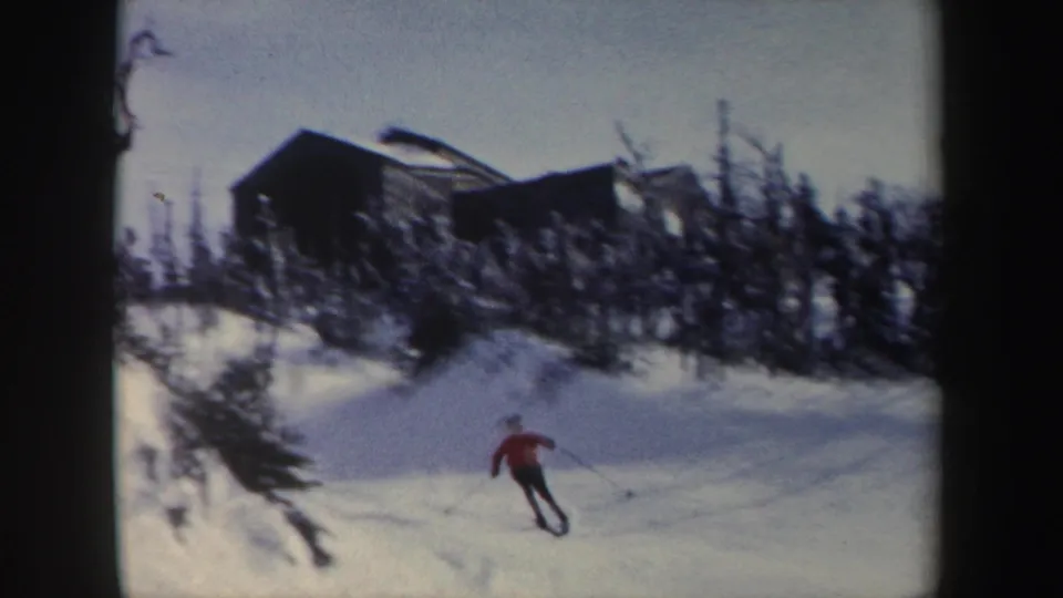 1962 New York 8mm frame showing a skier in a red jacket gliding downhill beneath a wooded ridge and cabin-like structure.