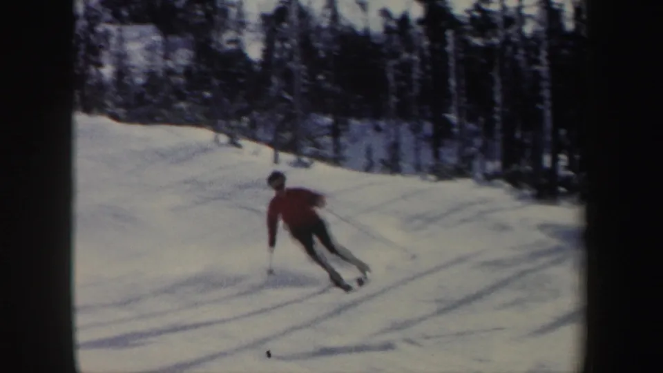 1962 New York 8mm frame showing a distant skier in a red jacket turning across a sunny snow-covered slope with trees behind.