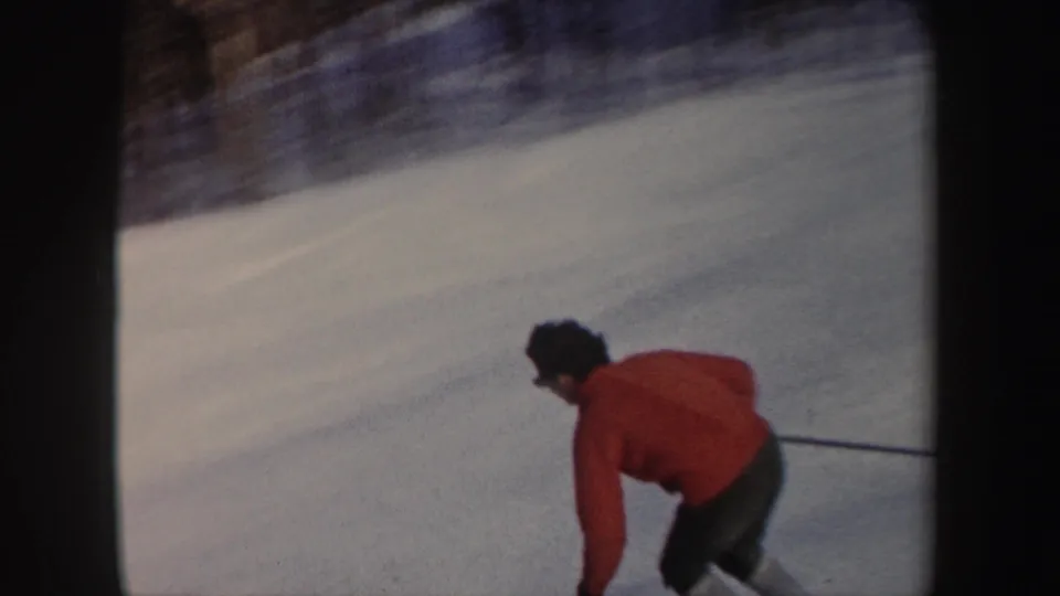 1962 New York home-movie frame of a skier in a red jacket leaning into a downhill run on a snow-covered slope.