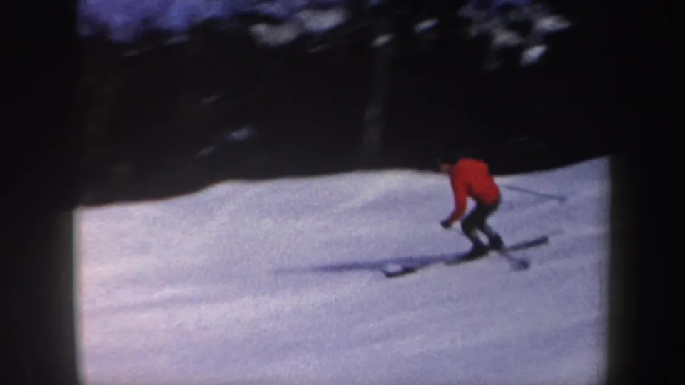 1962 New York 8mm frame with a skier in red leaning into a descent on a snowy slope below a dark treeline.