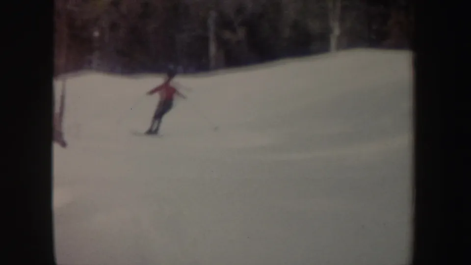 A skier in a red jacket glides across a snowy slope with a wooded background, captured in soft 1962 New York 8mm color.
