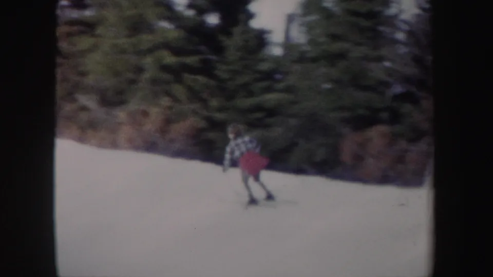 The skier continues descending the snowy slope, framed small against dark evergreen woods in 1962 New York footage.