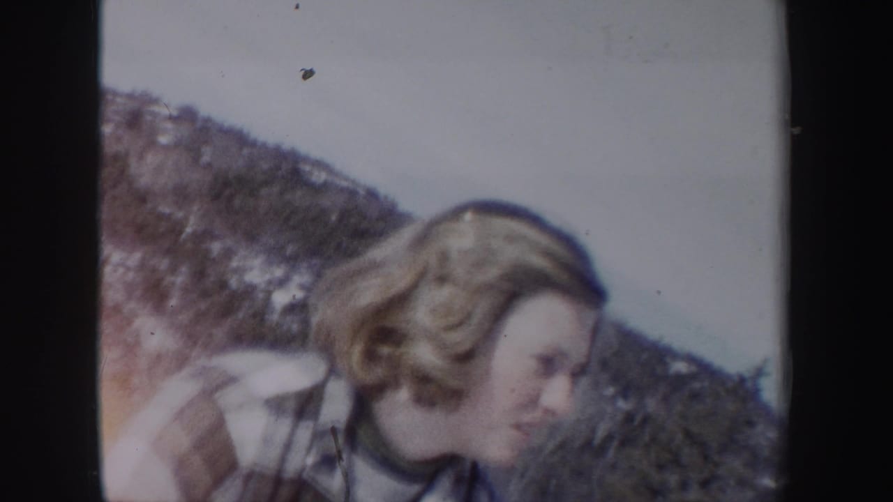 1962 New York home-movie frame of a woman in a plaid coat looking down while standing on a snowy mountain slope.