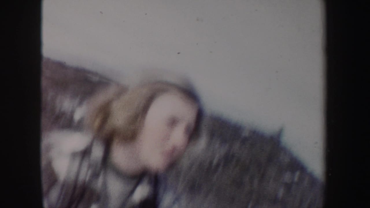 Soft 8mm frame from 1962 New York showing the woman blurred in the foreground against a snowy hillside.