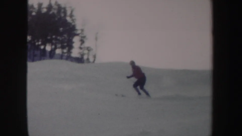 1962 New York 8mm frame showing a skier in a red top moving downhill across a snowy slope with trees in the background.