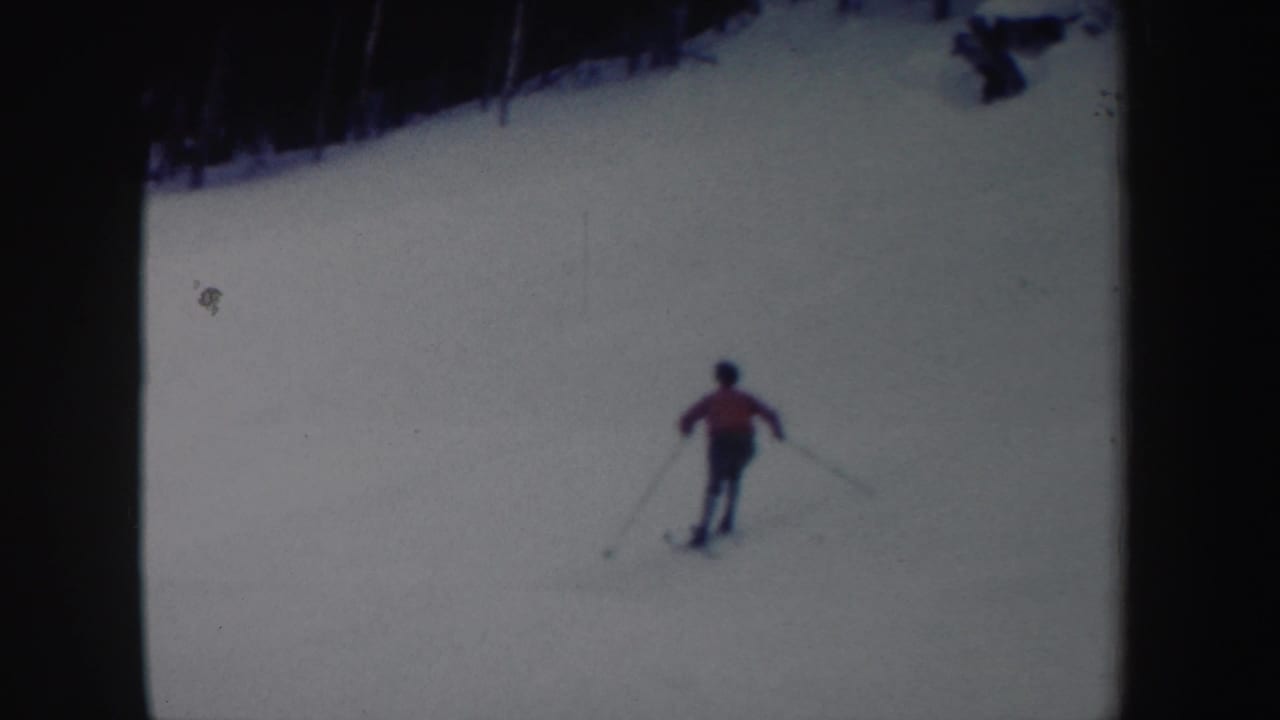 Skier in a red jacket descends a snowy slope near a dark treeline.