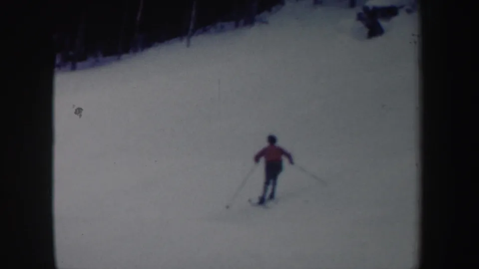 1962 New York 8mm frame of a skier in a red jacket moving downhill on a bright snowy slope with trees above.